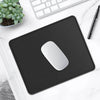A black non-slip mouse pad with a white mouse resting on a white marble surface next to a laptop keyboard and a pen.