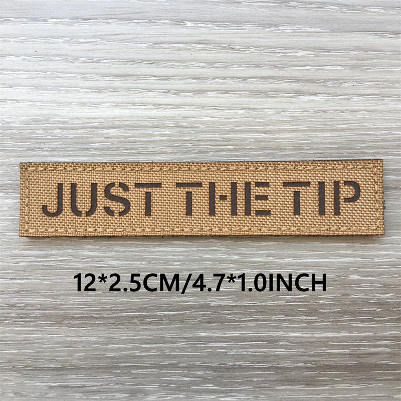 A rectangular tan woven patch with the text JUST THE TIP on a light gray wooden surface.