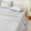 A light gray quilted bedspread with purple floral accents and a matching pillow on a white bed.
