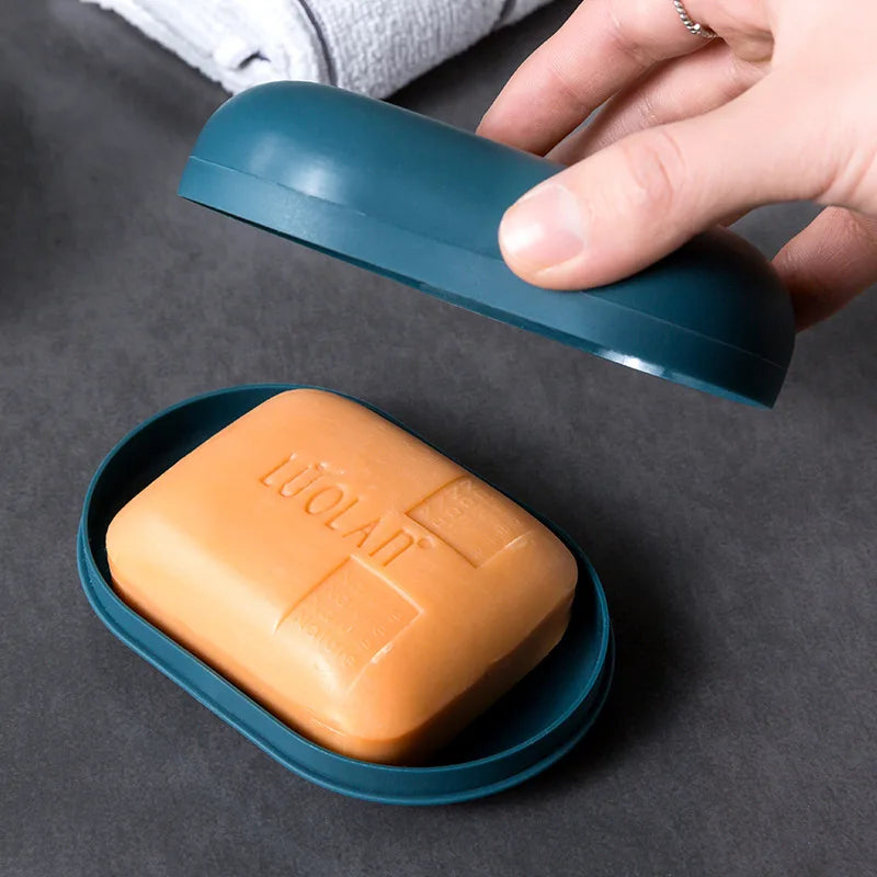 A hand holds a teal portable soap dish with a lid above an orange bar of soap in a matching dish.