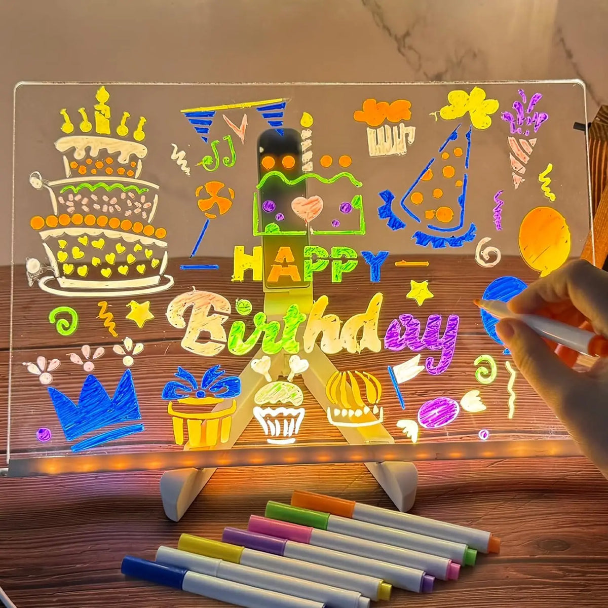 An LED drawing board illuminated by a night light features colorful drawings of a birthday cake and text on a transparent sur