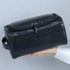 A black leather toiletry bag with a zipper is held in a person's hand against a light gray background.