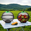 A black metal fruit basket with a mesh cover sits on a white table outdoors against a green landscape.