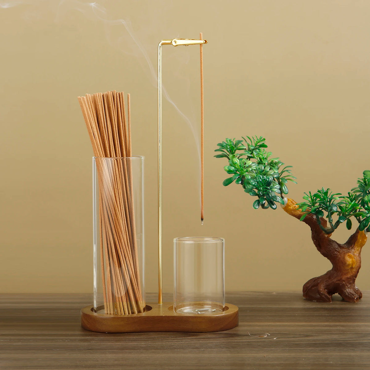 A tall glass incense holder with a wooden base and a thin metal rod emitting smoke, next to a small bonsai tree.