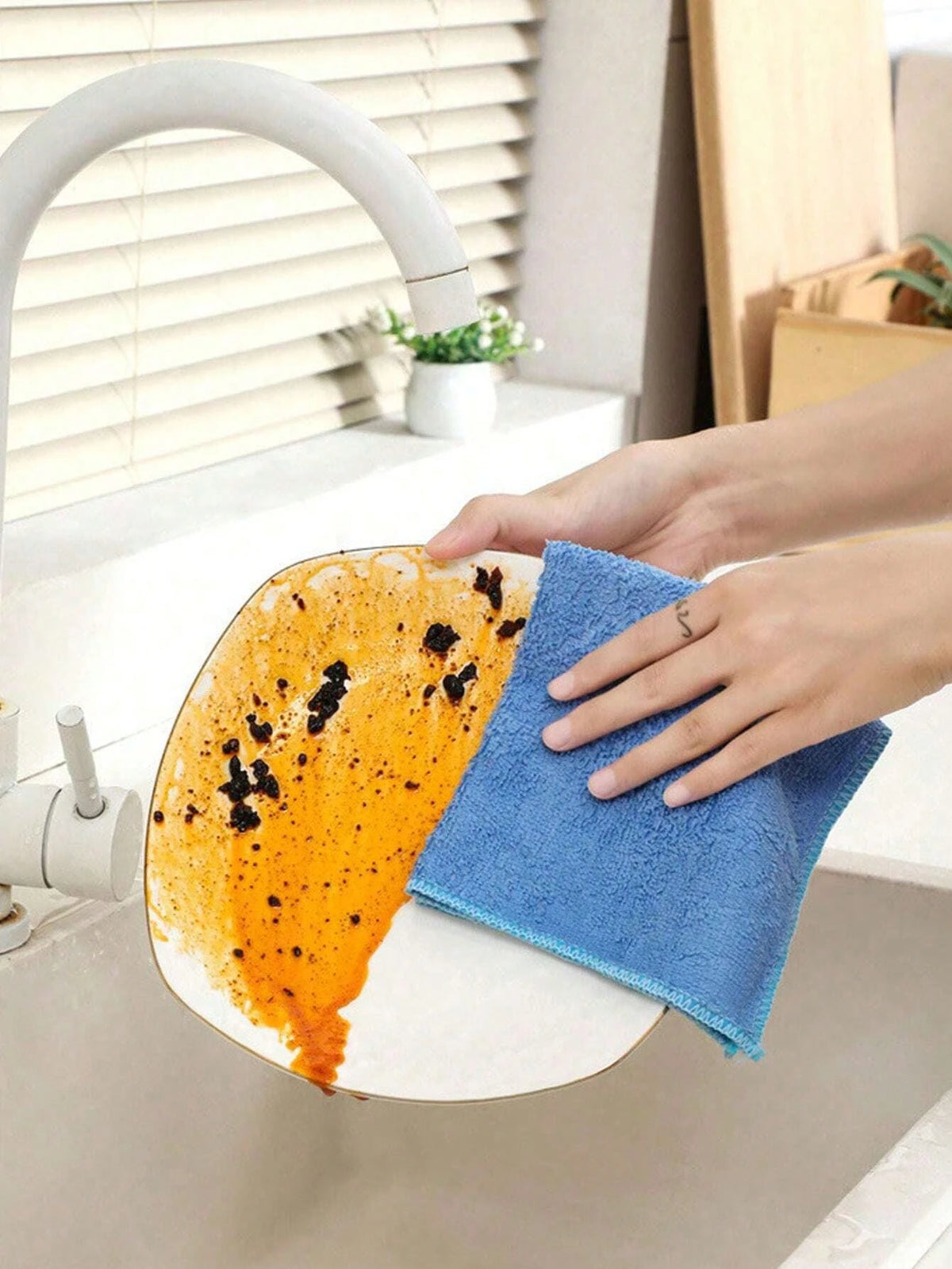 A hand is wiping an orange-colored citrus fruit with a blue microfiber cleaning cloth in a kitchen sink.