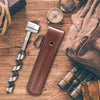 A brown leather holster for a manual wood auger drill, resting on a wooden surface with related tools.