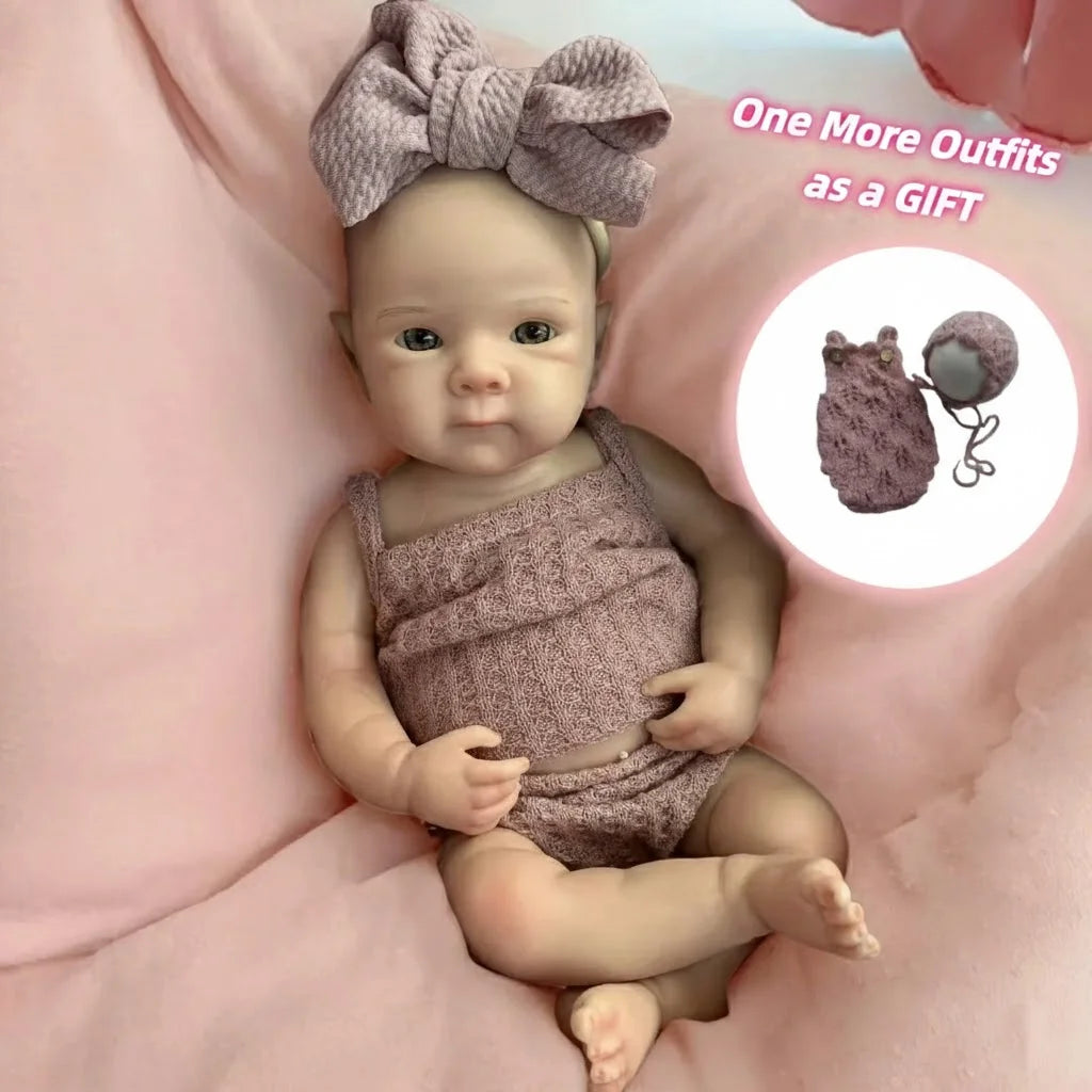A newborn silicone doll wearing a pink outfit and a bow, lying on pink fabric with a small accessory shown nearby.