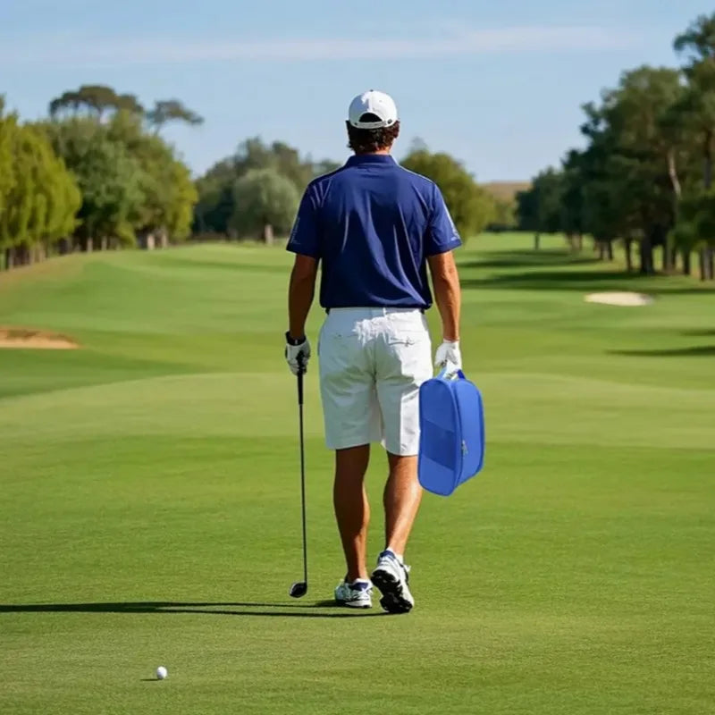 A man in a blue shirt and white shorts stands on a golf course, holding a golf club and carrying a blue shoe storage bag.