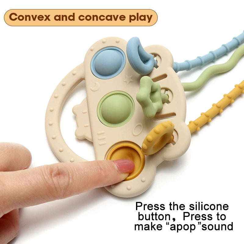 A hand holds a beige silicone bear teether with various colored textured parts and a pull string.