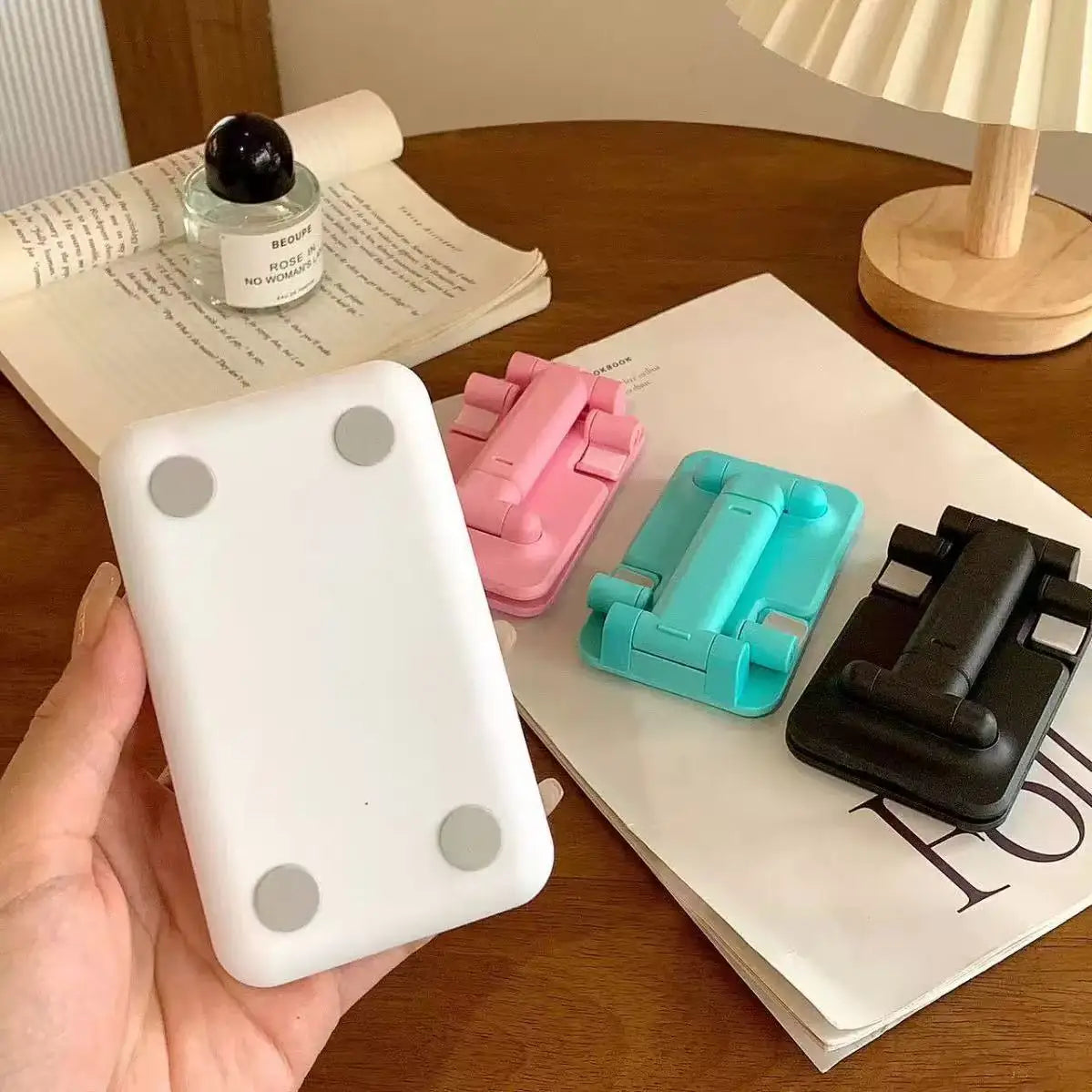 A white foldable desktop phone and tablet stand is held above a stack of papers on a wooden table.