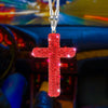 A red rhinestone cross pendant suspended by a silver chain, displayed in a car interior setting.
