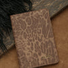 A brown leopard print passport holder with gold name embossing resting on a tan surface.