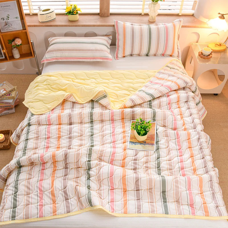 A quilted summer blanket in striped pink, green, and white fabric is displayed on a bed with a yellow throw.