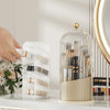 A clear glass rotating makeup brush holder with a gold lid and various makeup brushes displayed on a white surface.