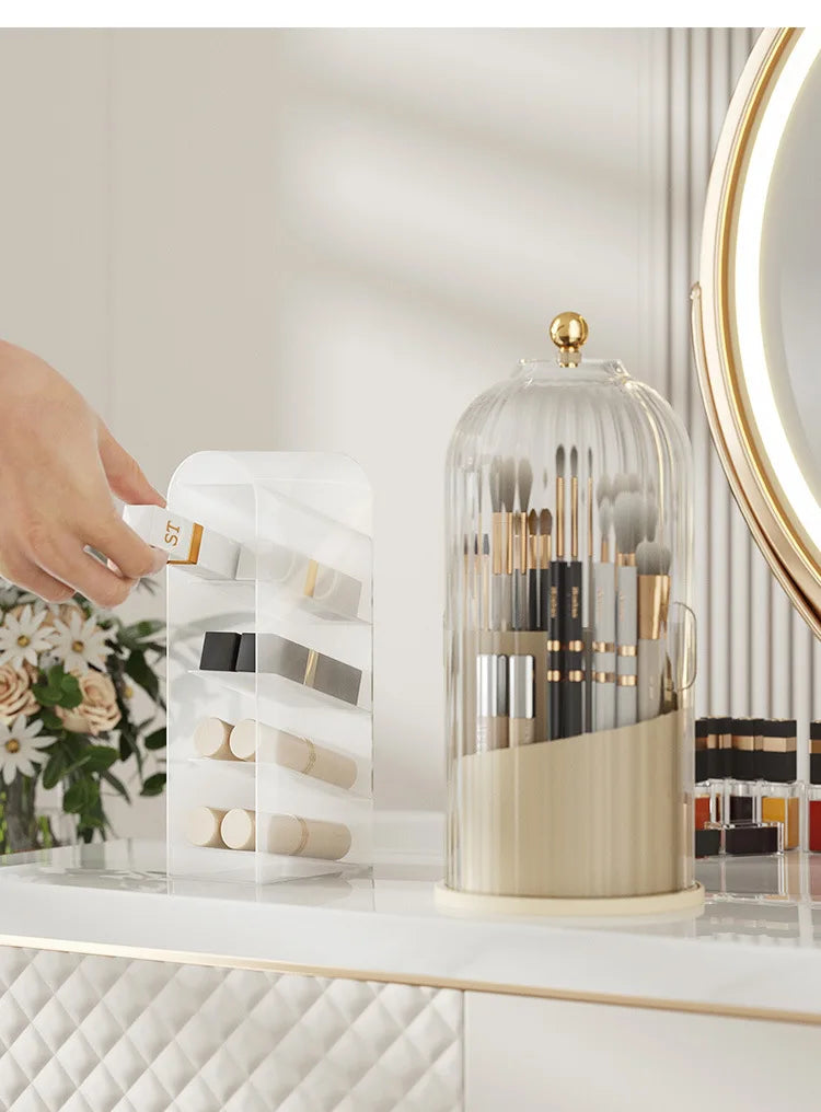 A clear glass rotating makeup brush holder with a gold lid and various makeup brushes displayed on a white surface.