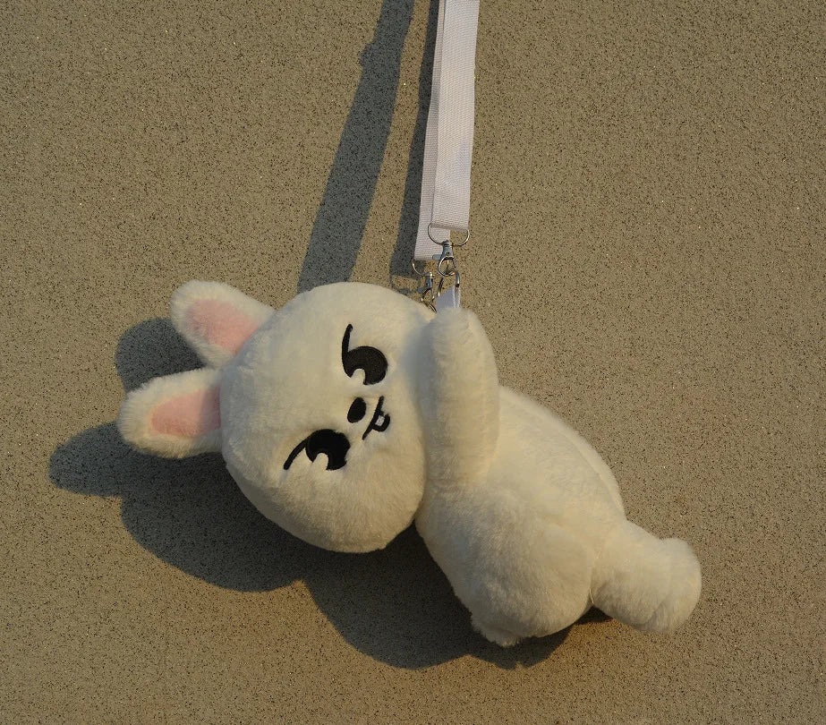 A white, fluffy cat-like plush doll with pink ears is hanging by a white strap against a beige background.