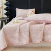 A pink floral quilted bedspread with a subtle plaid pattern and embroidered details is displayed on a bed.