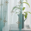 A sheer teal curtain decorated with delicate floral embroidery, featuring a green fern plant in a vase.