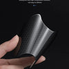 A hand holds a black carbon fiber door handle protector sticker, showing its texture and shape.