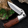 A folding pocket knife with a stainless steel blade and a walnut wood handle rests on a dark surface next to a piece of rock.