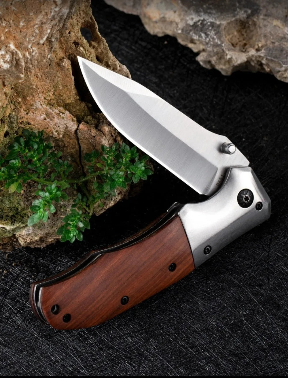 A folding pocket knife with a stainless steel blade and a walnut wood handle rests on a dark surface next to a piece of rock.