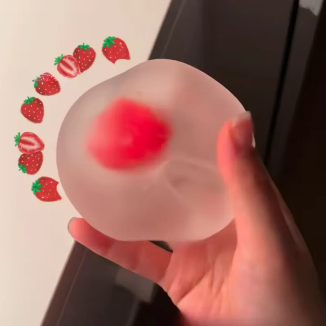 A hand holds a clear squishy toy with strawberry-shaped markers, showing a red strawberry being pressed into the silicone.