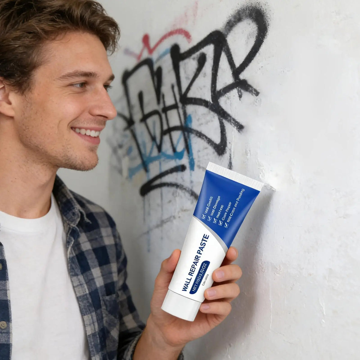 A man holds a tube of Wall Repair Paste for drywall repair against a white wall with black graffiti.