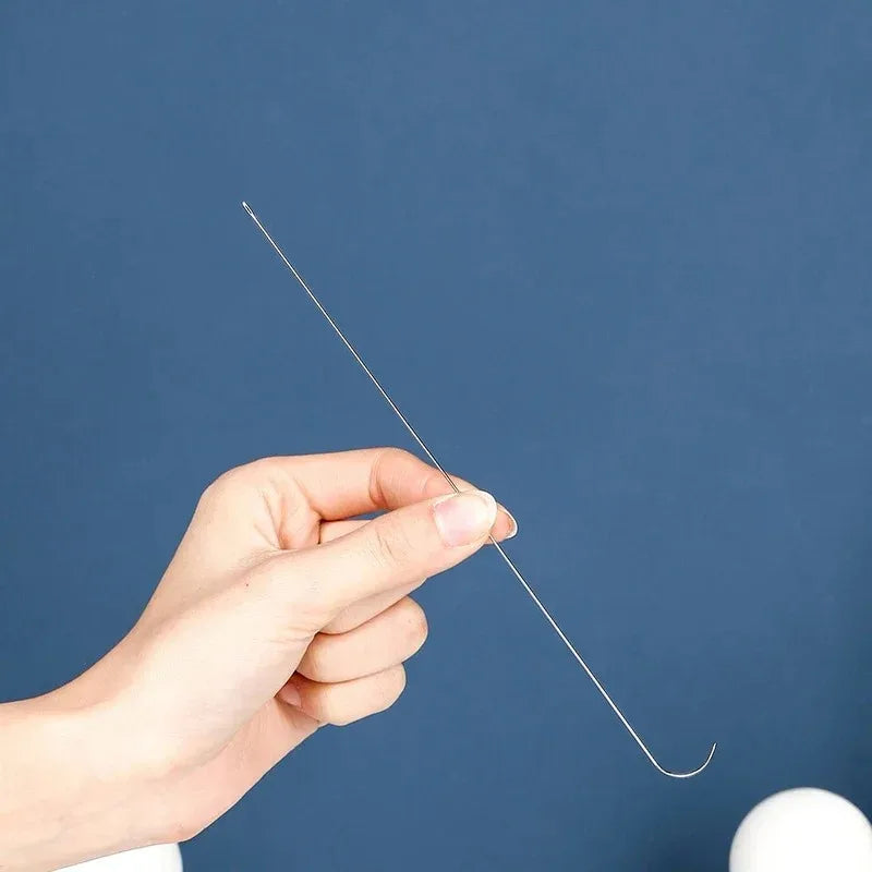 A hand is holding a thin, curved stainless steel beading needle against a dark blue background.