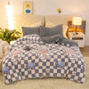 A gray and white checkered plush throw blanket is displayed on a light-colored rug next to a bed.