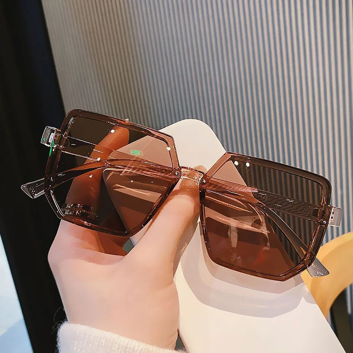Una mano sostiene unas gafas de sol cuadradas vintage marrones contra un fondo blanco.