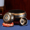 A handcrafted Tibetan singing bowl resting on a red cushion with its striker and cushion visible.