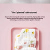 A pink tray displays a stack of white cotton muslin bandana bibs with colorful fruit and floral prints.