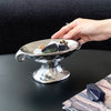 A hand holds a silver ceramic soap dish with natural elements on a dark surface.