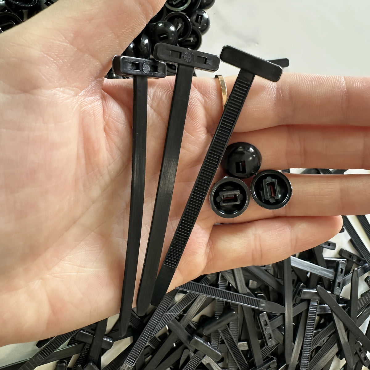 A hand holds several black self-locking nylon cable ties with push clips and a pile of similar ties.