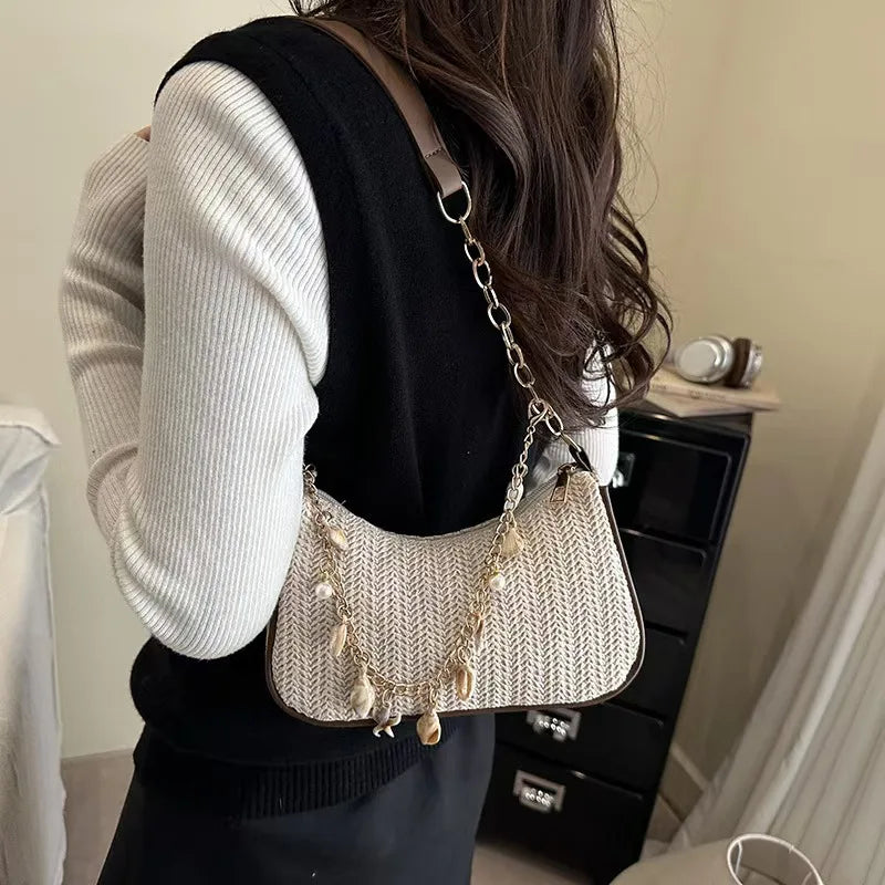 A woman is holding a beige woven straw shoulder bag with gold charms against a light background.