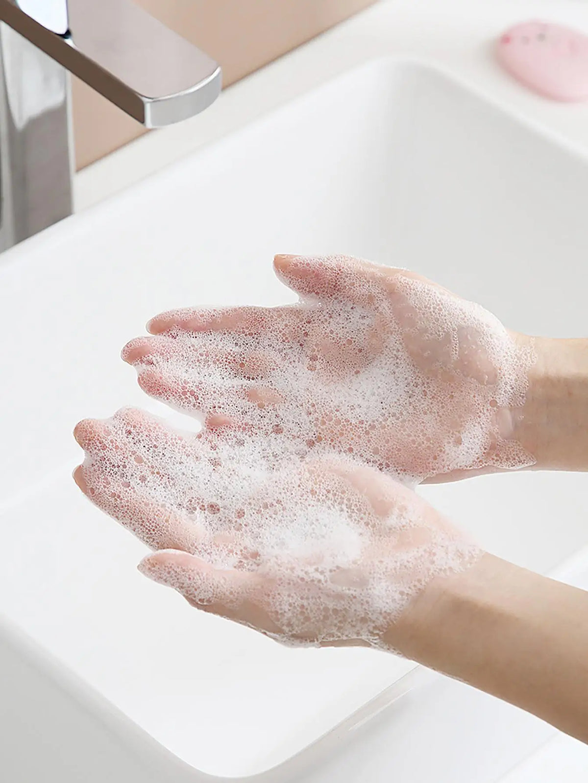 A person's hands are covered in white soap bubbles while washing them under a faucet.