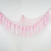 Pink bohemian handwoven corner toy storage net with sparkly tassels hanging on a white wall.