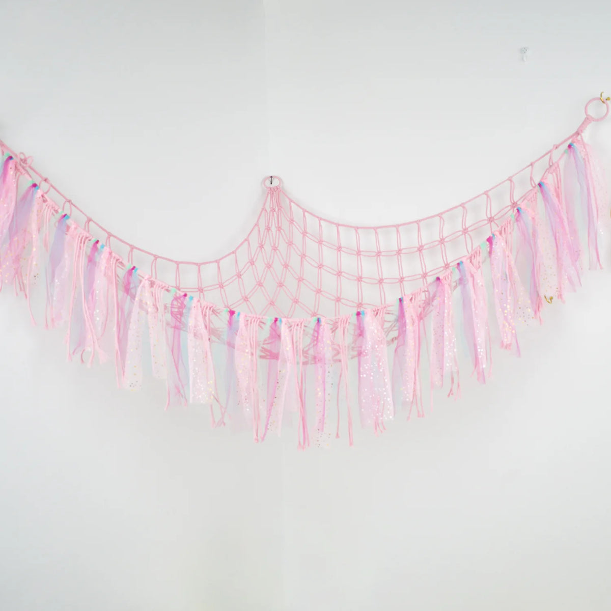 Pink bohemian handwoven corner toy storage net with sparkly tassels hanging on a white wall.