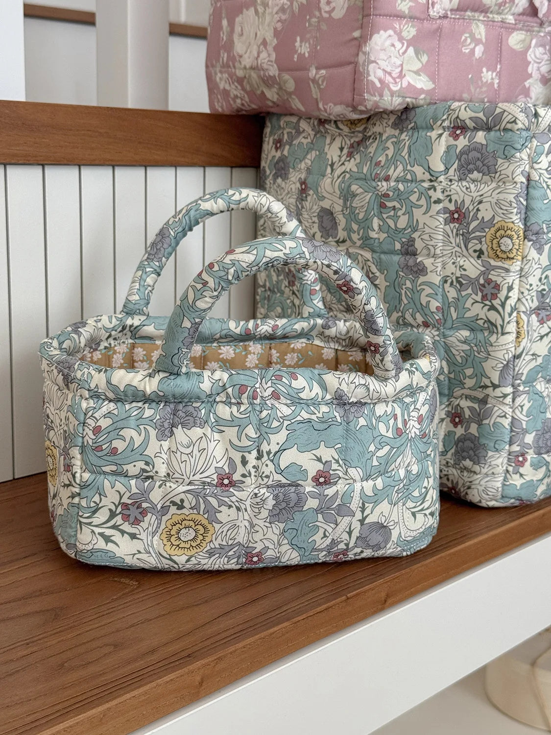 A quilted fabric storage basket in light blue and cream floral pattern sits on a wooden shelf.