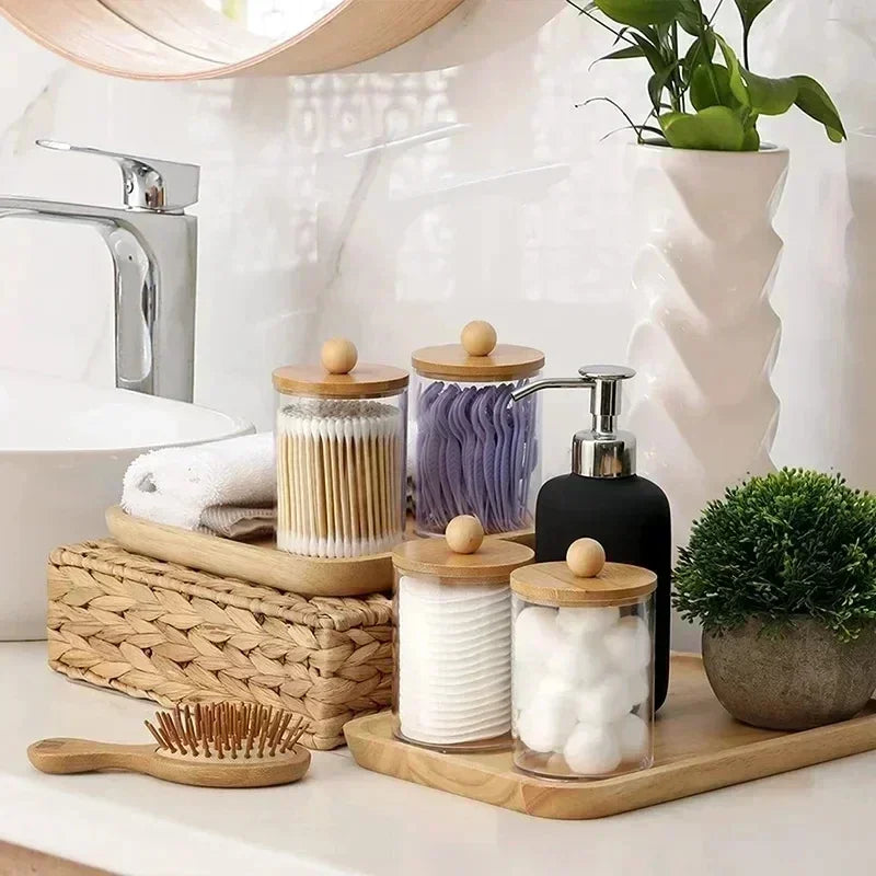 A set of clear apothecary jars with wooden lids and a pump dispenser for bathroom organization on a white countertop.