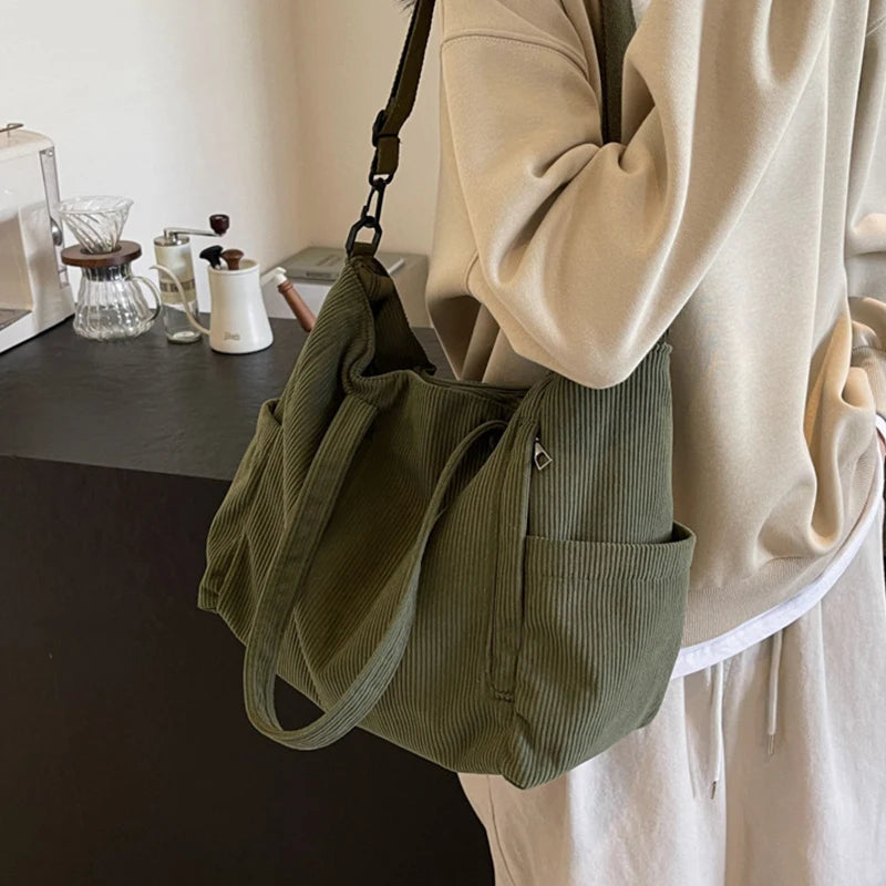 A beige top is holding a olive green corduroy tote bag with an adjustable strap.