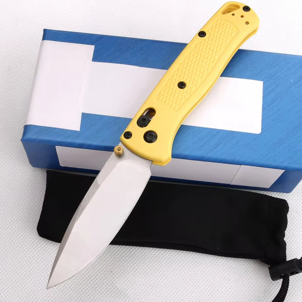 A yellow folding knife with a stainless steel blade is shown resting on a blue box and a black sheath.