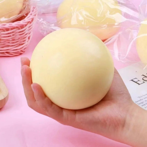 A hand holds a smooth, pale yellow stress relief ball packaged in clear plastic.