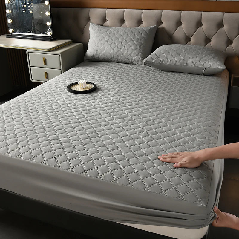 A gray quilted waterproof mattress protector is shown on a bed, with a decorative pillow and a small candle on the surface.