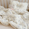 A white muslin baby blanket with a light blue floral pattern is folded on a light pink surface next to a white pillow.