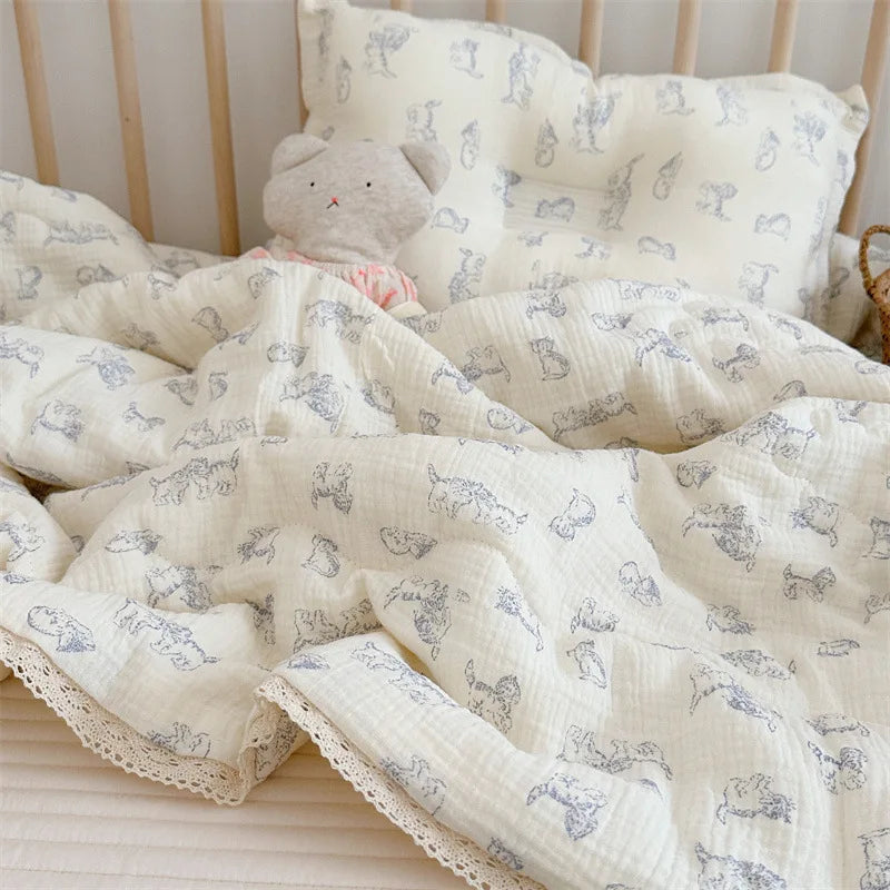 A white muslin baby blanket with a light blue floral pattern is folded on a light pink surface next to a white pillow.