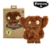 A brown, fuzzy Fuggler Ugly Monster plush toy with gold eyes and funny teeth displayed next to its packaging.