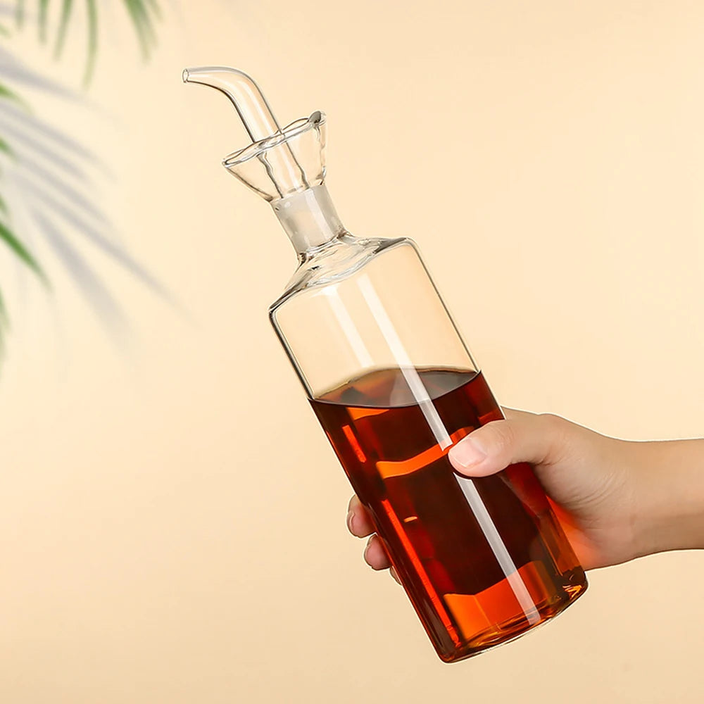 A hand holds a glass oil and vinegar dispenser with a spout, showing the amber liquid inside.
