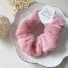A pink plush coil hair tie is displayed on a white plate with a tag, next to a light-colored woven object.