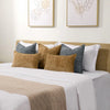A collection of decorative cushion covers in various shades of gray and gold resting on a white bed with a beige throw blanke